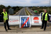 Els portaveus del moviment veïnal 'Mai més peatges', Llorenç Navarro i Enric Lange, mostren les pancartes de l'entitat des de dalt d'un pont sobre l'autopista AP-7 a l'altrua de l'Aldea