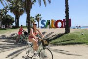 Diversos turistes en bicicleta que passen per davant d'un rètol amb lletres de colors on es llegeix 'Salou'.