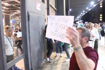 Un home amb un cartell en què ofereix quatre places al cotxe per anar al Maresme, davant de Sants de Barcelona, mentre un viatger des de dins de l'estació observa l'exterior, aquest dilluns a la nit després de l'apagada general.