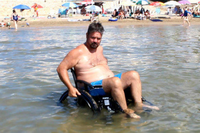 Pla general d'un home prenent el bany a la platja amb una cadira de rodes adaptada.