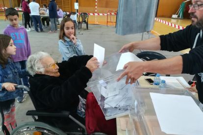 L'Amèlia Guardià de l'Arboç als seu 107 anys exercint el seu dret a vot, acompanyada de la besnéta, que agafa la cadira i el seu besnét.