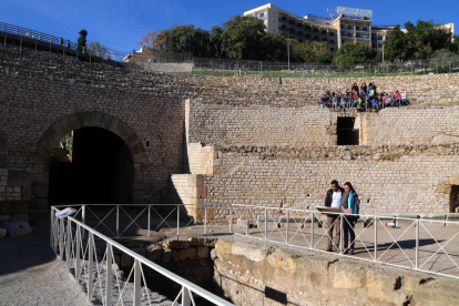Dos turistes visitant l'amfiteatre romà de Tarragona amb un grup d'escolars assegut a la graderia.