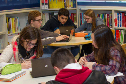 Pla general de sis alumnes treballant en equips de tres en taules rodones amb ordinadors, en una biblioteca de l'escola pia.