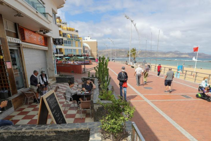 Diverses persones disfrutande un cafè en una terrassa mentre unes altres caminen pel passeig de la platja de les Pedreres, a Las Palmas de Gran Canaria.