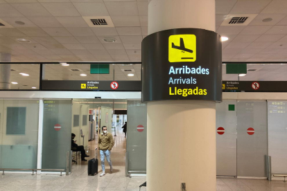 Un senyal de la zona d'Arribades amb un passatger arrossegant una maleta de fons a l'Aeroport del Prat