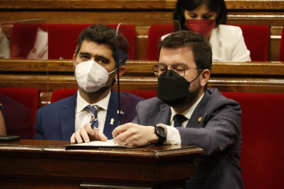 El president de la Generalitat, Pere Aragonès, escrivint en un paper, i al costat el vicepresident Jordi Puigneró als escons de l'hemicicle del Parlament.