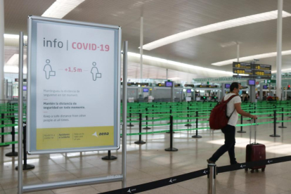 Un cartell informatiu de la covid-19 i d'un passatger arrossegant una maleta amb el control de seguretat de la T1 de l'Aeroport del Prat.