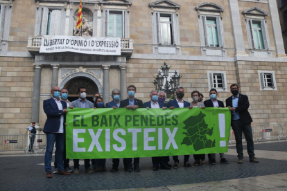 Plano abierto de los 14 alcaldes del Baix Penedès mostrando una pancarta delante del Palau de la Generalitat, en la plaza Sant Jaume de Barcelona, donde se lee 'El Baix Penedès existe'.