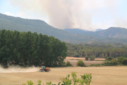 Un tractor llaurant un camp de cereals i al fons les flames de l'incendi que crema les comarques de la Conca de Barberà i l'Anoia.