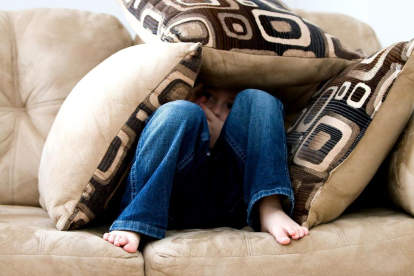 Imagen de recurso de un niño escondido entre cojines|almohadas en un sofá.