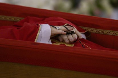 El papa descansa sobre un féretro de madera forrado de terciopelo rojo con una casulla púrpura y una mitra blanca y a las manos lleva un rosario.