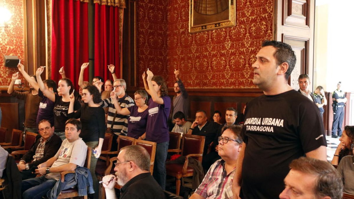 Un agent de la Guàrdia Urbana en primer pla i diversos activistes alçant guixos de color blanc al fons, al ple d'aquest dilluns.