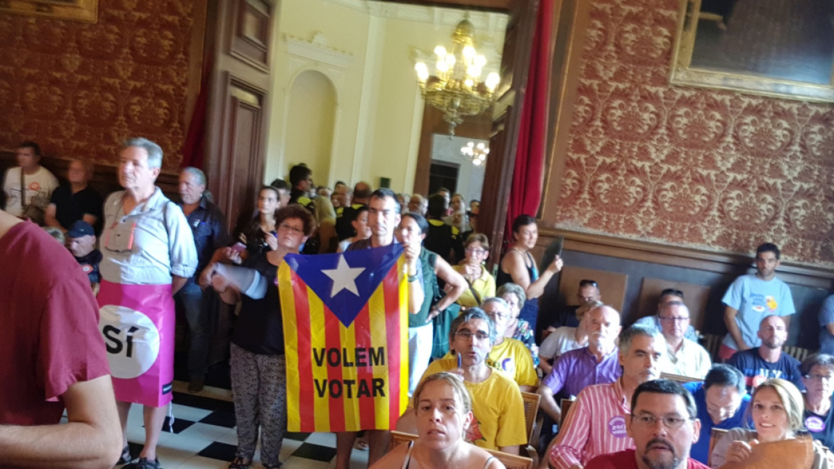 Imagen de asistentes al pleno con banderas en qué se puede leer 'sí' y 'queremos votar'.