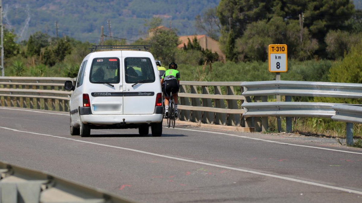 Pla general del punt quilomètric 8 de la T-310 entre Riudoms i Montbrió, amb un grup de ciclistes circulant mentre un cotxe els intenta avançar hores després d'un accident mortal amb dos ciclistes en el mateix punt.
