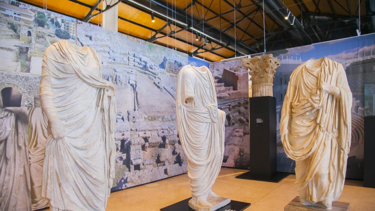 Tres estatuas, la de la izquierda de una mujer, y, como fondo, fotografías del Anfiteatro y del Teatro de la ciudad romana de Tarraco.