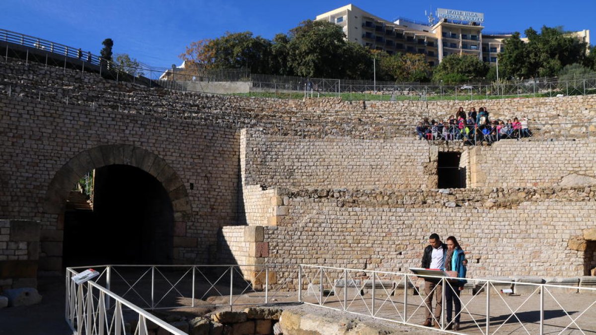 Dos turistas visitando el anfiteatro romano de Tarragona con un grupo de escolares sentado en la gradería.