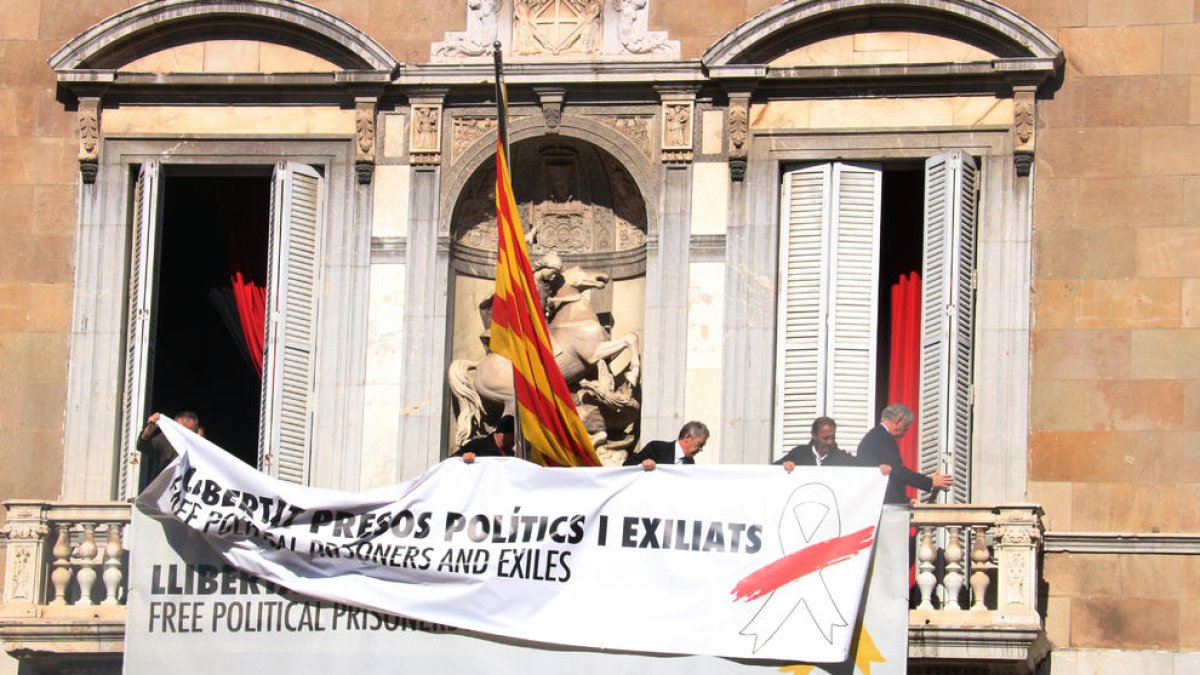 Treballadors del Govern afegeixen una pancarta damunt l'anterior, ara amb el llaç blanc i una franja vermella, al balcó del Palau de la Generalitat.