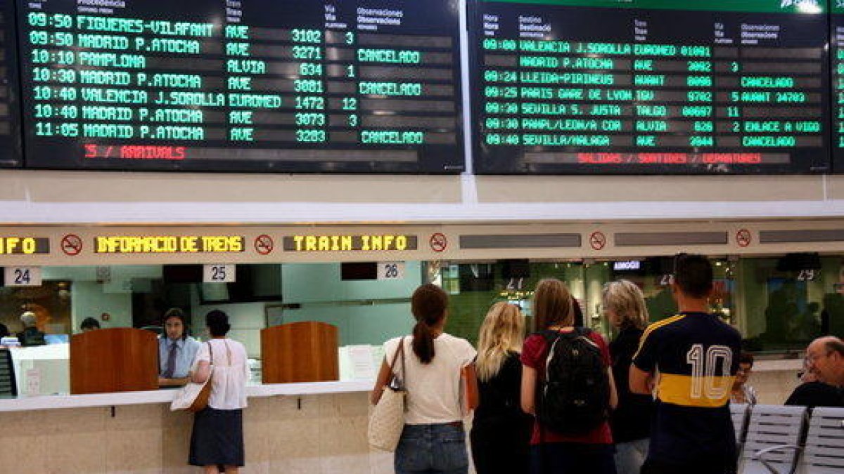 Imagen de usuarios de Renfe haciendo cola en la taquilla de información bajo el cartel de salidas y llegadas a la estación de Sants.