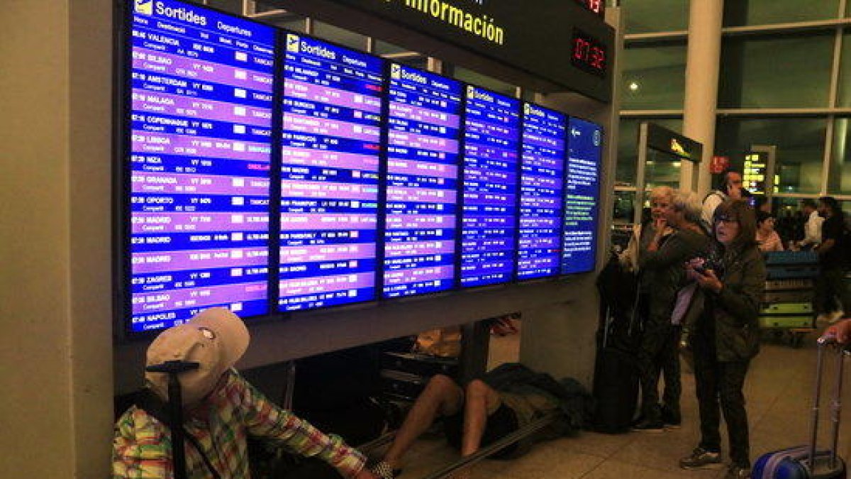 Pasajeros consultando los paneles informativos de salidas mientras un hombre duerme debajo, en la terminal 1 del aeropuerto del Prat, al día siguiente del bloqueo por parte de miles de manifestantes después de conocerse la sentencia del 1-O.