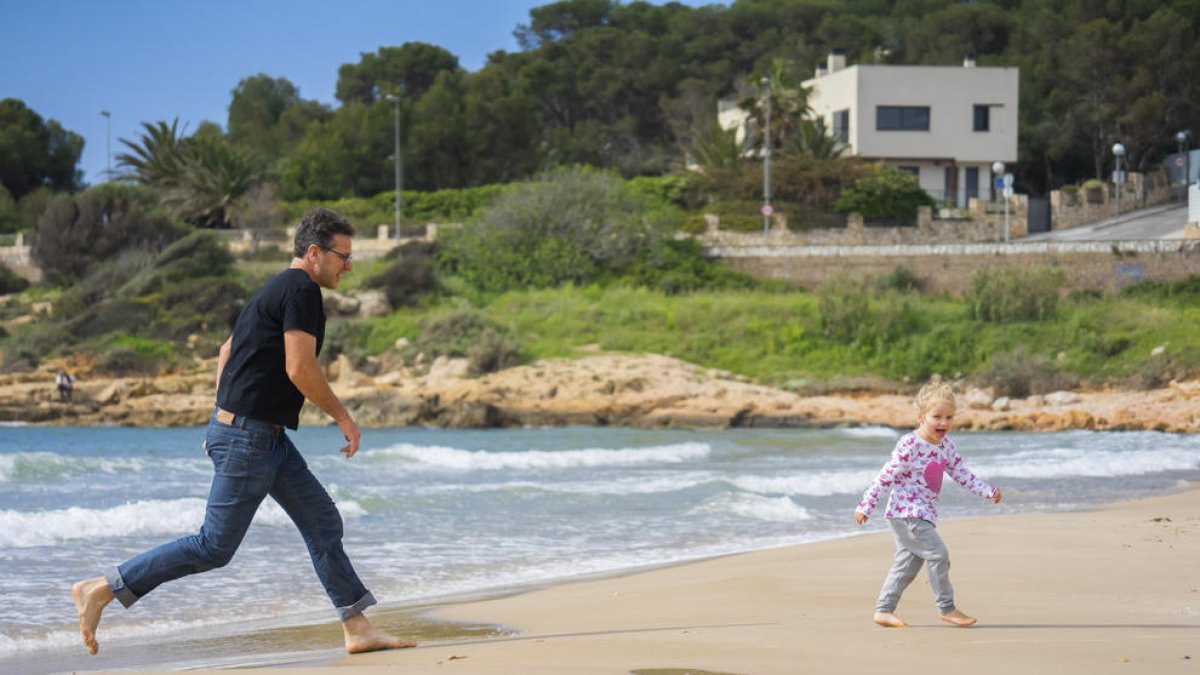 Un pare jugant i corrent amb la seva filla per la sorra de la platja de l'Arrabassada, espai escollit per moltes famílies per passejar i sortir una estona.