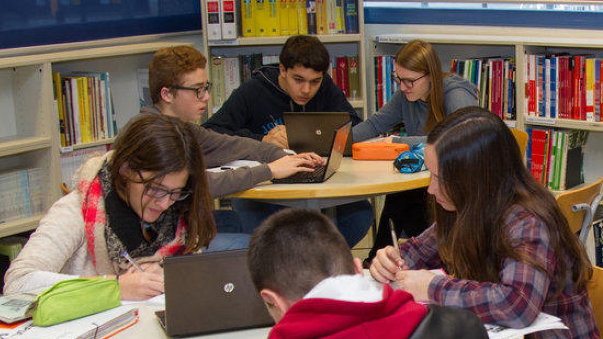 Plano general de seis alumnos trabajando en equipos de tres en mesas redondas con ordenadores, en una biblioteca de la escuela pia.