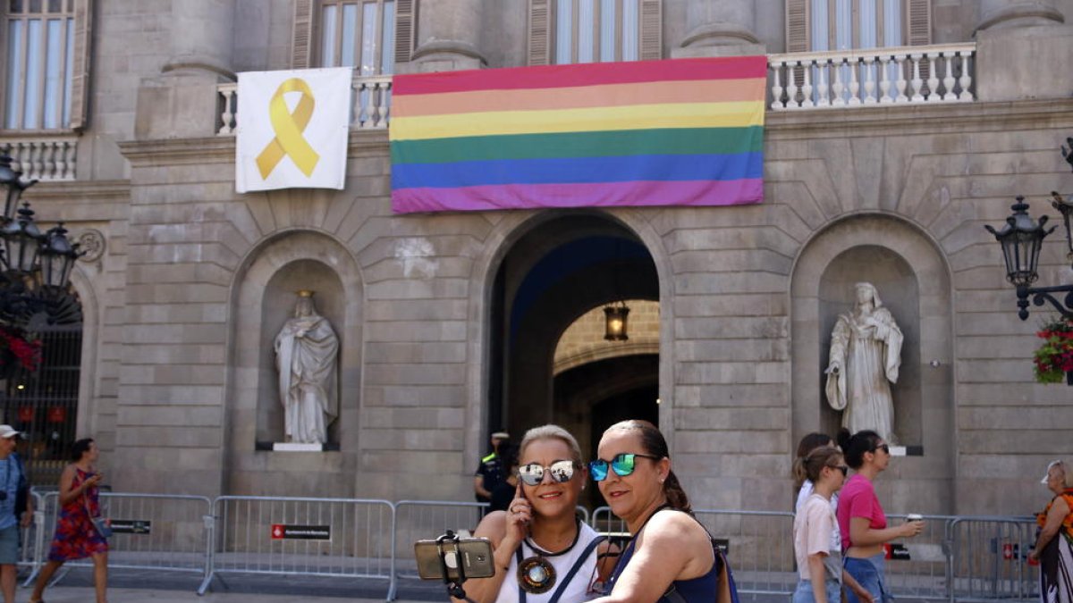 Dues dones fotografiant-se davant de la façana de l'Ajuntament de Barcelona amb la bandera LGTBI de l'Arc de Sant Martí penjada.