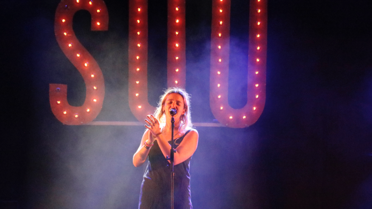 Pla mitjà de la cantant Suu amb el nom del grup de fons durant el seu concert al Festival Acústica de Figueres el 29 d'agost de 2019