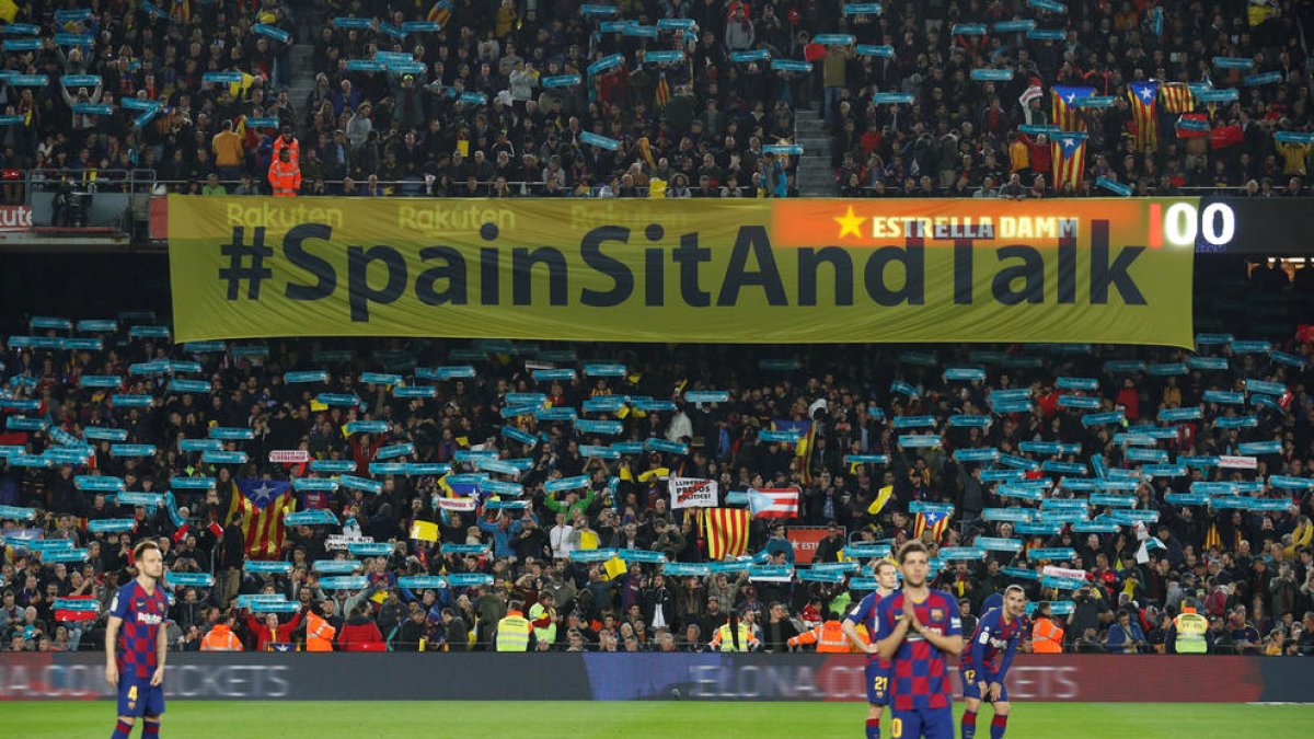 Cartell de Tsunami Democràtic amb el lema 'Spain, sit and talk!' a l'inici del clàssic al Camp Nou darrere dels jugadors.