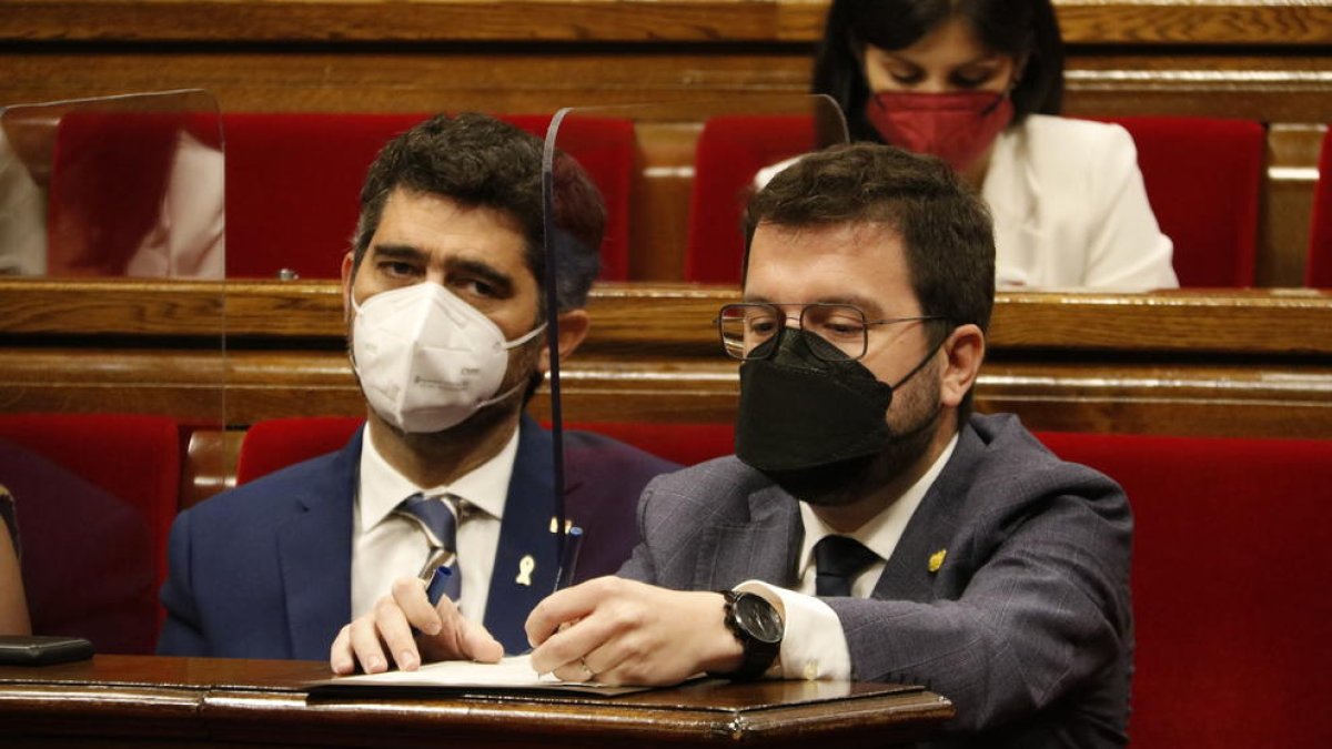 El presidente de la Generalitat, Pere Aragonès, escribiendo en un papel, y al lado el vicepresidente Jordi Puigneró en los escaños del hemiciclo del Parlamento.