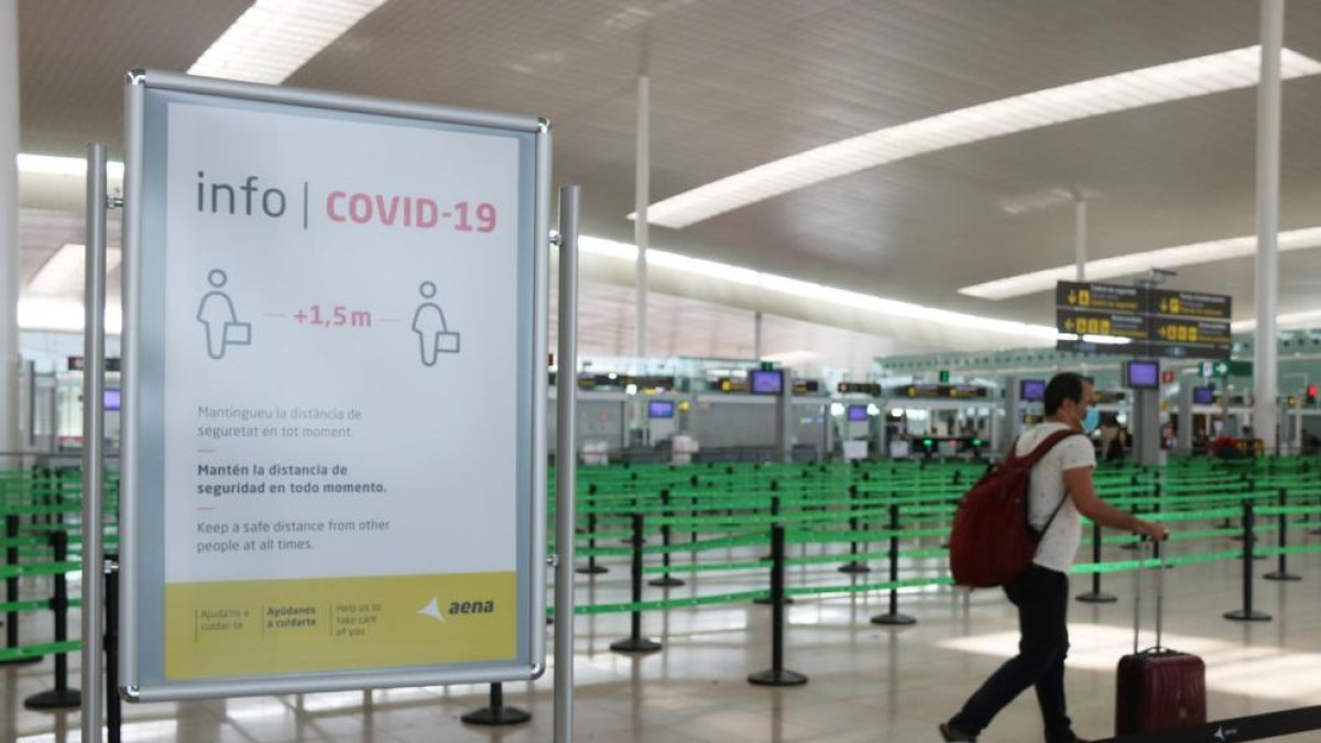 Un cartell informatiu de la covid-19 i d'un passatger arrossegant una maleta amb el control de seguretat de la T1 de l'Aeroport del Prat.