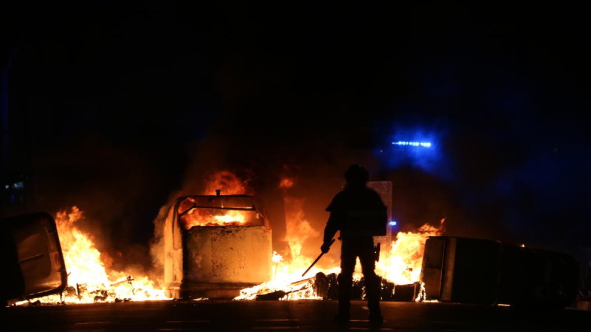 Silueta d'un agent dels antiavalots dels Mossos d'Esquadra davant de diversos contenidors cremats durant les protestes.