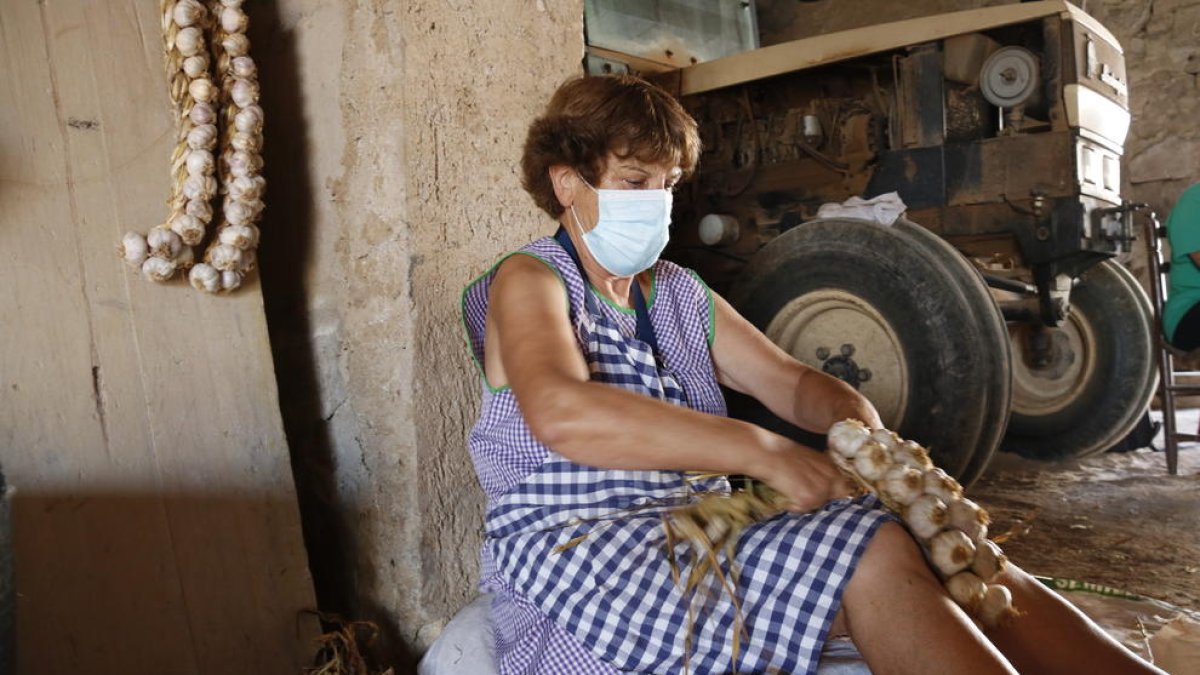 Una dona asseguda al portal de casa seva fent forcs d'alls a Belltall (Conca de Barberà), amb un tractor al fons.