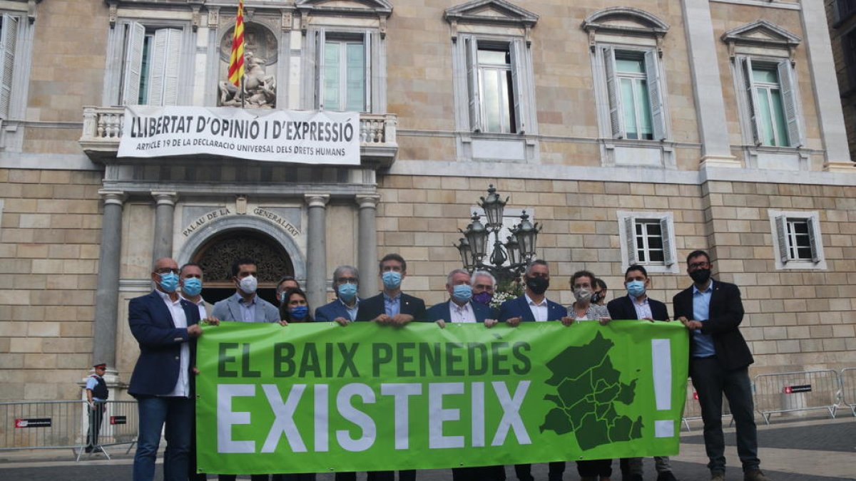 Plano abierto de los 14 alcaldes del Baix Penedès mostrando una pancarta delante del Palau de la Generalitat, en la plaza Sant Jaume de Barcelona, donde se lee 'El Baix Penedès existe'.