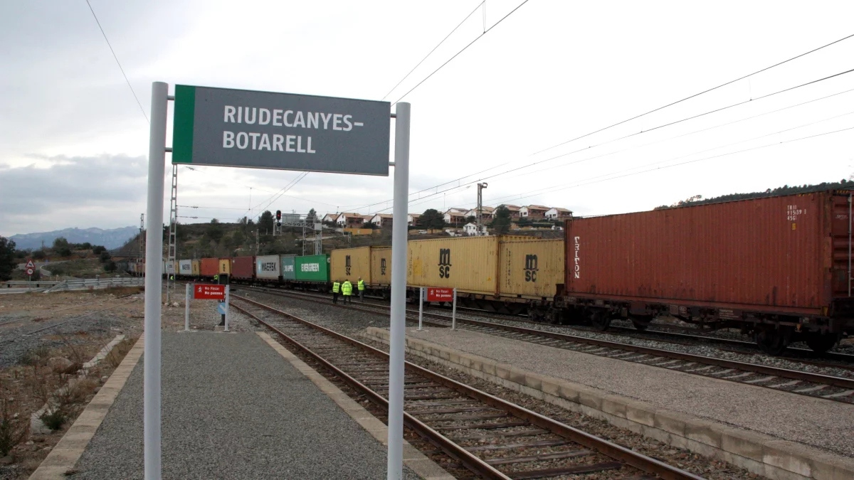 Imatge d'arxiu d'un cartell de l'estació de Riudecanyes-Botarell, en primer terme, amb el tren de mercaderies descarrilat i operaris al fons