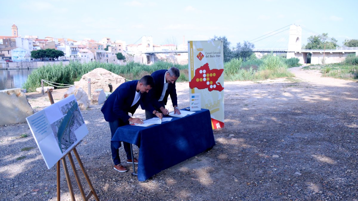 El president del Consell del Baix Ebre, Antoni Gilabert, i l'alcalde de l'Aldea, Xavier Royo, signen la cessió dels terrenys amb el pont penjant d'Amposta i el tram de l'enllaç de la via verda, al fons.