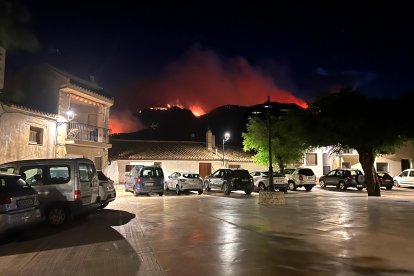 Cotxes aparcats a la plaça major de Paüls amb el foc cremant al fons.
