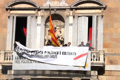 Trabajadores del Gobierno añaden una pancarta encima del anterior, ahora con el lazo blanco y una franja roja, en el balcón del Palau de la Generalitat.