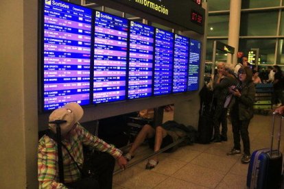 Pasajeros consultando los paneles informativos de salidas mientras un hombre duerme debajo, en la terminal 1 del aeropuerto del Prat, al día siguiente del bloqueo por parte de miles de manifestantes después de conocerse la sentencia del 1-O.