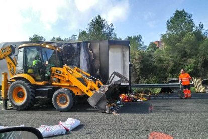 Imatge del camió incendiat, amb les fruites i verdures al terra i operaris treballant en la retirada de la mercaderia.