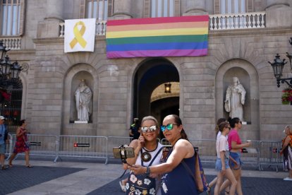 Dos mujeres fotografiándose delante de la fachada del Ayuntamiento de Barcelona con la bandera LGTBI del Arco Iris colgada.