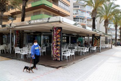 Plano abierto de una mujer paseando un perro cerca de una terraza del paseo de Salou prácticamente sin clientes