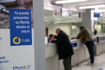 Un cartel en la sede de la Agencia Tributaria en Cataluña con indicaciones sobre la presentación de la declaración de la renta y con contribuyentes haciendo consultas de fondo,