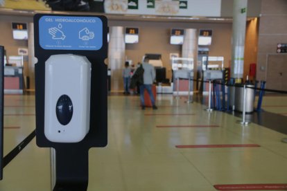 Un dispensador de hielo|gel hidroalcòholic en el aeropuerto de Reus con una cola de pasajeros en el fondo facturando.