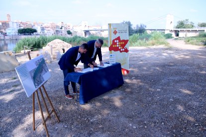 El president del Consell del Baix Ebre, Antoni Gilabert, i l'alcalde de l'Aldea, Xavier Royo, signen la cessió dels terrenys amb el pont penjant d'Amposta i el tram de l'enllaç de la via verda, al fons.