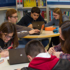 Pla general de sis alumnes treballant en equips de tres en taules rodones amb ordinadors, en una biblioteca de l'escola pia.