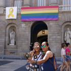 Dos mujeres fotografiándose delante de la fachada del Ayuntamiento de Barcelona con la bandera LGTBI del Arco Iris colgada.