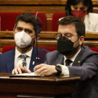 El presidente de la Generalitat, Pere Aragonès, escribiendo en un papel, y al lado el vicepresidente Jordi Puigneró en los escaños del hemiciclo del Parlamento.