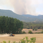 Un tractor llaurant un camp de cereals i al fons les flames de l'incendi que crema les comarques de la Conca de Barberà i l'Anoia.