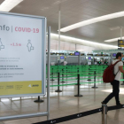 Imatge d'un cartell informatiu de la covid-19 i un passatger arrossegant una maleta a la T1 de l'Aeroport del Prat.
