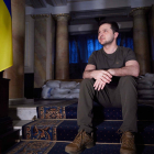El presidente de Ucrania, Volodímir Zelenski, sentado en una escalera, con sacos de protección detrás y una bandera del país al lado.